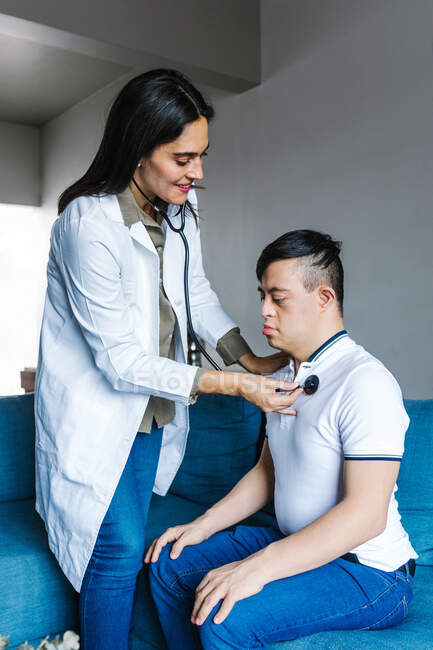 Веселая женщина-медик осматривает латинского подростка с синдромом Дауна со стетоскопом во время приема на дому — стоковое фото