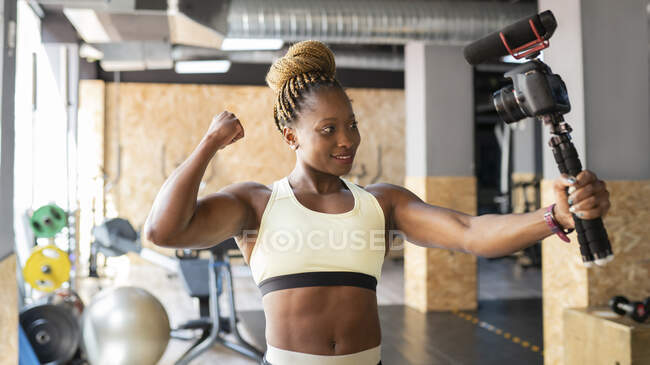 Jovem se encaixa atleta afro-americana demonstrando músculos enquanto grava vídeo na câmera fotográfica no ginásio — Fotografia de Stock