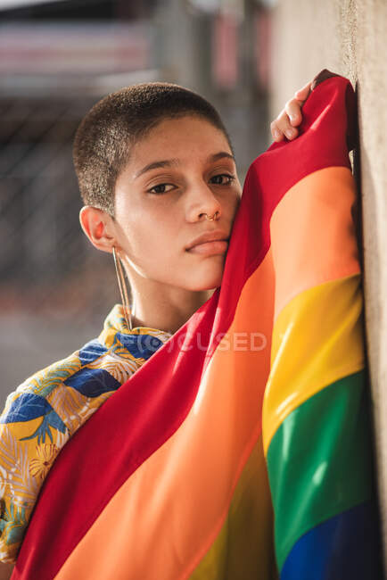 Joven mujer étnica soñadora con bandera colorida y pelo corto mirando a la cámara contra la pared sobre fondo borroso - foto de stock