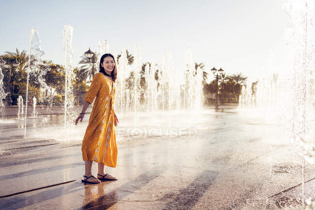 Vue latérale complète du corps de la femme gaie en robe élégante profitant de l'eau douce de la fontaine Emirates Palace à Abu Dhabi tout en passant des vacances d'été dans les Émirats — Photo de stock