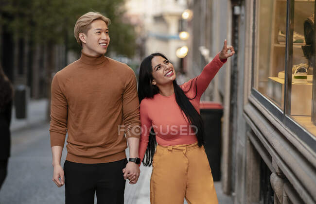 Веселая многорасовая пара, держащаяся за руки и идущая по улице, указывая на витрину модного магазина — стоковое фото
