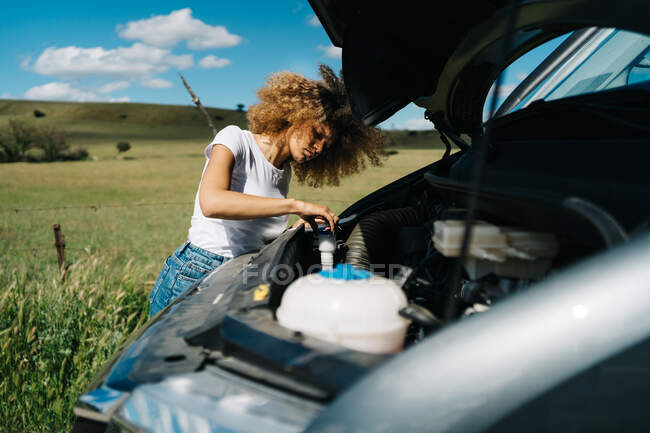 Side view of traveling female looking under opened hood of camper van while having problems during trip through summer nature — Stock Photo