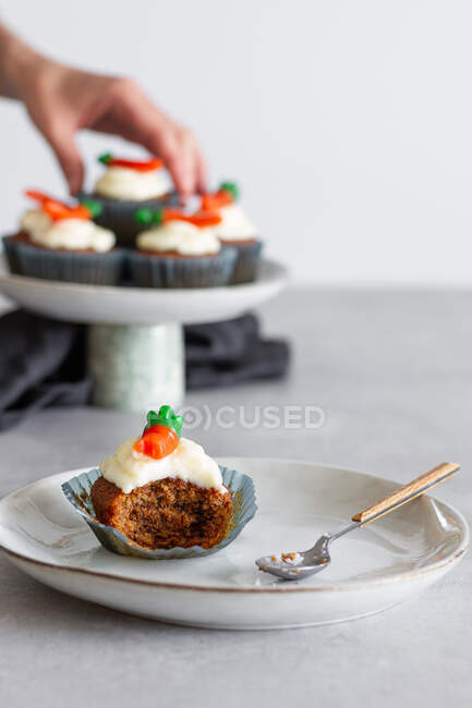 Cupcake à la carotte à moitié mangé avec de la crème sur l'assiette contre la personne floue traitant avec le dessert — Photo de stock