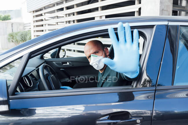 Side view of serious man using a protective mask driving car during quarantine time making top gesture with hand looking at camera — Stock Photo