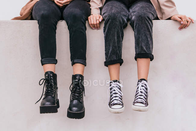 Mulheres irreconhecíveis em jeans elegantes e calçados sentados na parede cinza na rua da cidade — Fotografia de Stock