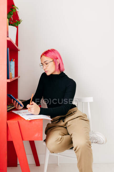 Боковой вид молодой креативной женщины-дизайнера с розовыми волосами в повседневной одежде и очках с помощью смартфона и рисования эскизов во время работы на рабочем столе дома — стоковое фото