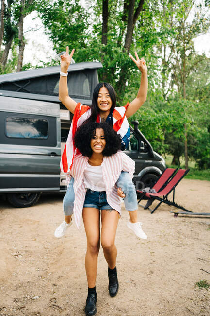Весела молода афроамериканська жінка, яка їздить на багатій азійській подружці з американським прапором, насолоджуючись літніми пригодами біля фургона-кемпера в лісі. — стокове фото
