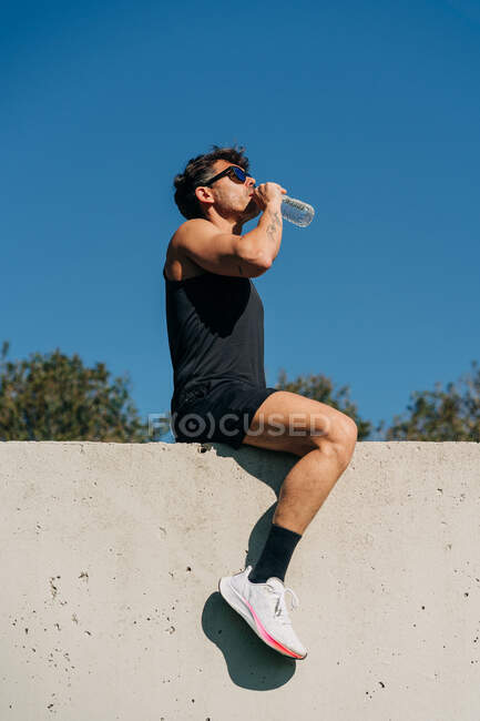 Вид збоку спортсмена в активному одязі, що п'є акваріум з пляшки після тренування проти дерев на сонячному світлі — стокове фото