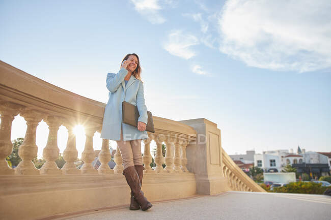 Mujer feliz con abrigo azul apoyada en valla de piedra con teléfono celular y carpeta en las manos en la calle de la ciudad en un día soleado - foto de stock