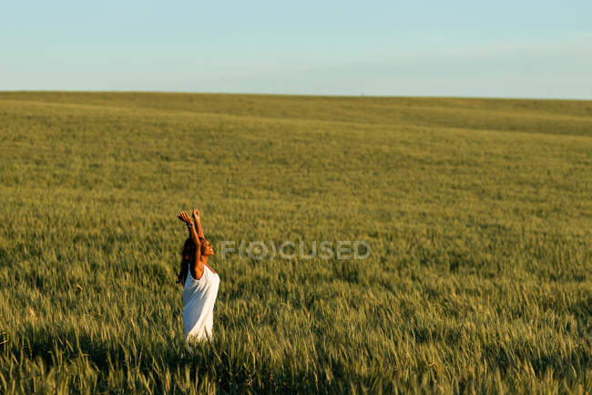 Giovane signora nera in bianco abito estivo passeggiando sul campo di grano verde, mentre guardando in alto di giorno sotto il cielo blu — Foto stock