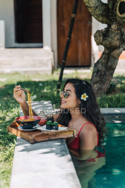 Vista lateral de la alegre viajera femenina en traje de baño contra la piscina con deliciosa pasta asiática cocinada entre palillos a la luz del sol - foto de stock