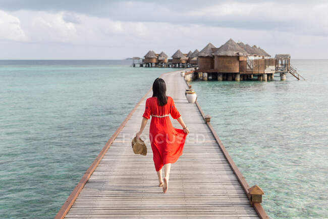 Обратный вид женщины в красном платье, гуляющей босиком по тропическому пляжному проспекту, расслабляющей на Мальдивах — стоковое фото
