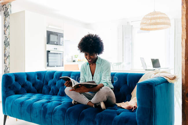 Joven hembra negra en atuendo casual con auriculares sentados en un acogedor sofá azul en casa y leyendo la revista mientras disfruta del tiempo libre en casa - foto de stock