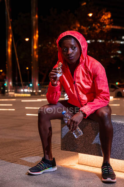 Young African American athletic woman having snack while sitting on street at night — Stock Photo