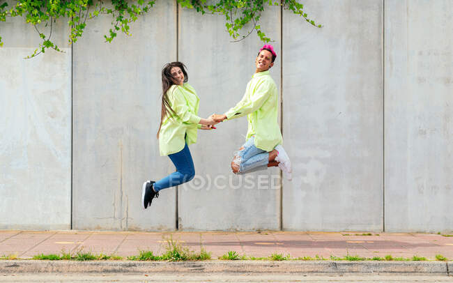 Vista lateral do casal animado de mãos dadas e pulando acima do solo enquanto se diverte e desfruta de liberdade no verão na cidade — Fotografia de Stock