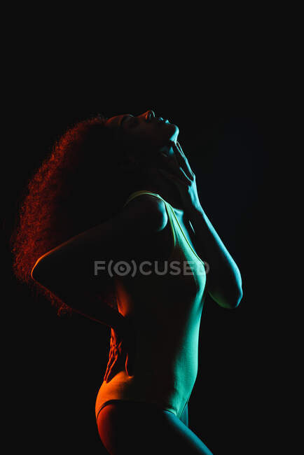 Бічний вид на молоду афро-американську жінку з кучерявим волоссям в нижній білизні з рукою на талії дотик підборіддя з закритими очима на чорному тлі студії — стокове фото