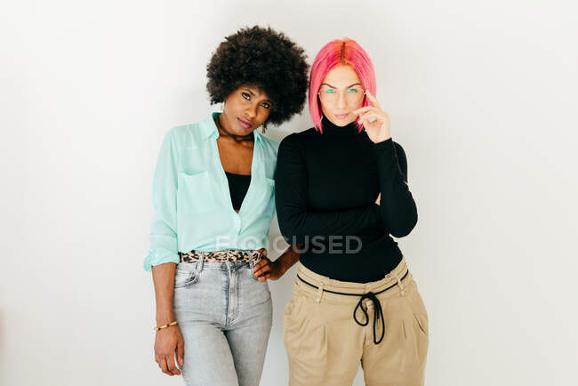 Весела молода рожева жінка та афроамериканська дівчина в стильному вбранні стоять разом на білому фоні. — стокове фото