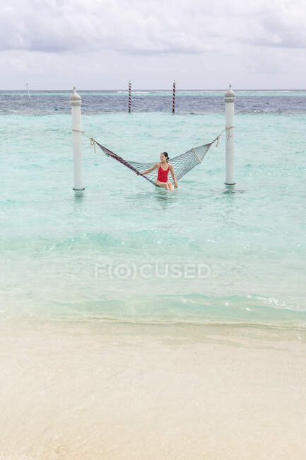 Mulher de fato de banho vermelho sentada na rede balançar sobre a linha de surf oceano relaxante em Maldivas no dia nublado — Fotografia de Stock