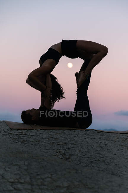 Вид сбоку на силуэт гибкой женщины, делающей спину и балансирующей на ногах мужчины во время сеанса акройоги на фоне закатного неба с луной — стоковое фото