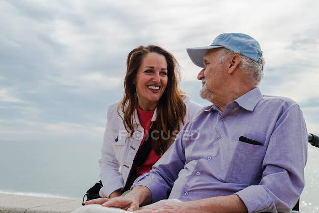Contenuto figlia adulta e padre anziano in sedia a rotelle agghiacciante su argine contro il mare insieme in estate — Foto stock