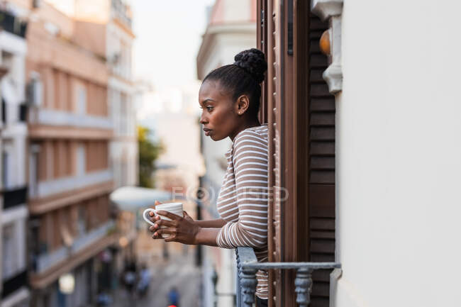 Vista laterale della donna afroamericana riflettente con tazza di bevanda calda guardando lontano dal balcone in città — Foto stock