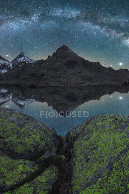 Paesaggio notturno mozzafiato di aspre montagne rocciose con neve vicino al lago calmo con superficie liscia dell'acqua che riflette il cielo con la Via Lattea lucida — Foto stock
