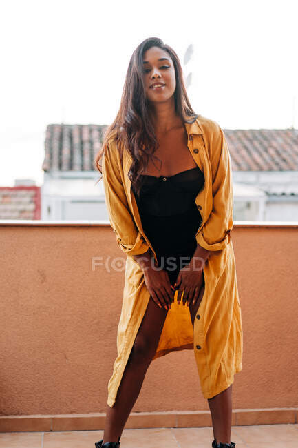 Frohe junge hispanische Frau in gelbem Baumwollmantel, die auf dem Balkon lächelt und in die Kamera blickt — Stockfoto