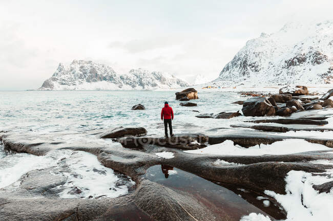 Visão traseira de ma irreconhecível admirando bela paisagem do mar frio com água salpicando em rochas perto da costa gelada e nevada perto de montanhas no dia de inverno cinza nas Ilhas Lofoten, Noruega — Fotografia de Stock