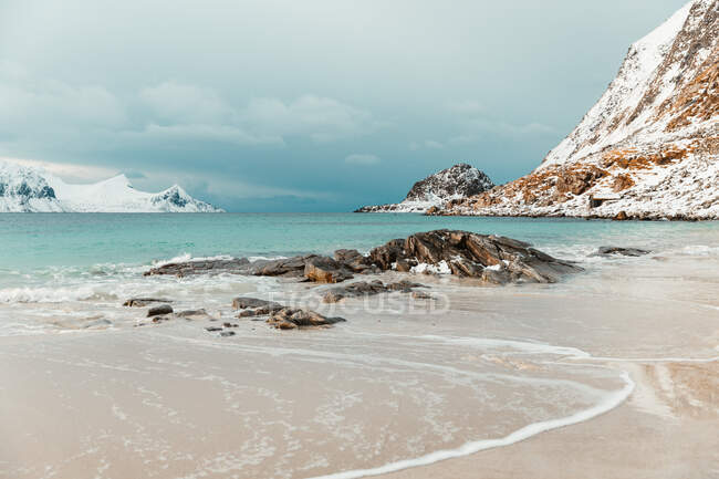Холодні морські хвилі котилися на березі біля засніжених гір проти хмарного неба в зимовий день на Лофотенських островах (Норвегія). — стокове фото