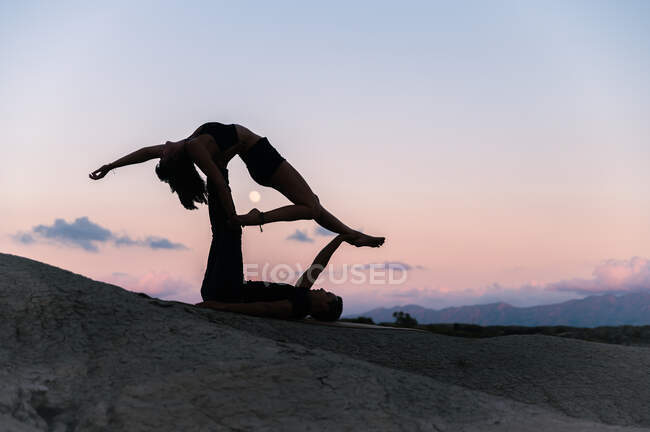 Vista lateral do casal gracioso fazendo acroyga no fundo do céu por do sol nas montanhas — Fotografia de Stock
