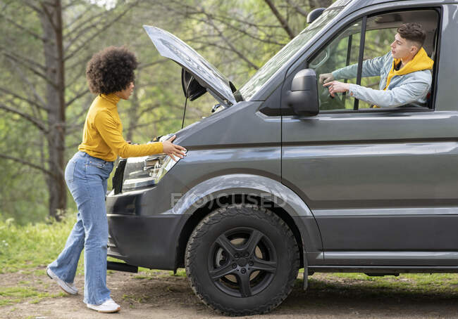 Vue latérale d'une femme noire avec une coiffure afro écoutant les instructions du petit ami et vérifiant le moteur de la camionnette tout en essayant de réparer le véhicule pendant le voyage à travers les bois — Photo de stock