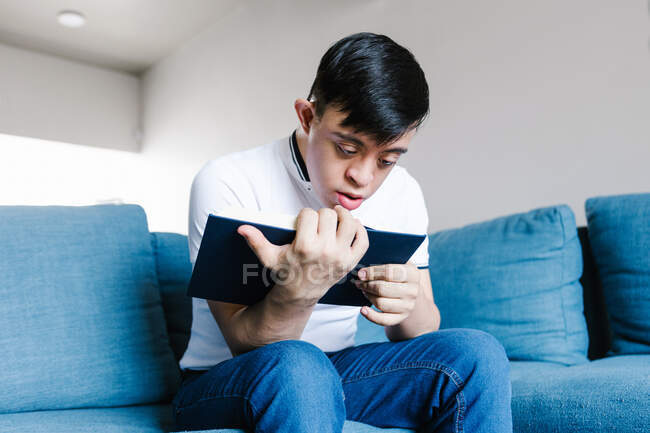 Baixo ângulo de adolescente étnico com síndrome de Down lendo livro interessante enquanto sentado no sofá na sala de estar em casa — Fotografia de Stock