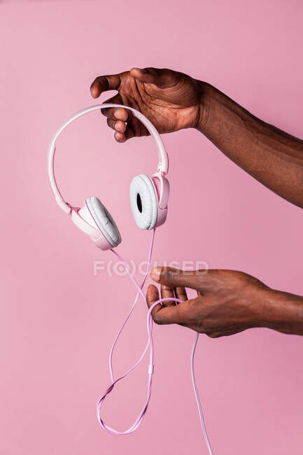 Mano de hombre negro anónimo sosteniendo auriculares rosados contra fondo de color a juego - foto de stock