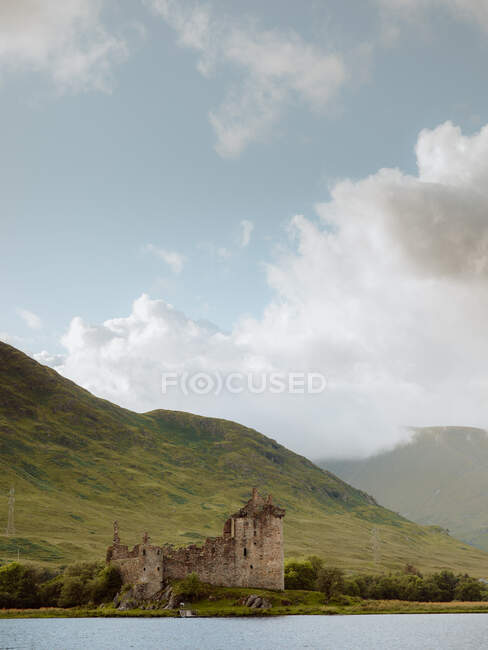 Поврежденный старый замок, расположенный на берегу спокойного озера против травянистых холмов на сельской местности в пасмурный день в замке Килчурн, Великобритания — стоковое фото