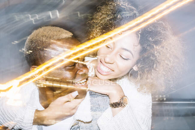 Alegre jovem afro-americano masculino dando passeio de piggyback para namorada feliz com cabelo encaracolado na roupa da moda perto de luzes congeladas — Fotografia de Stock