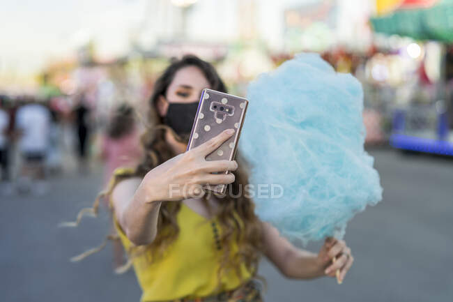 Mujer en máscara protectora y con azúcar de algodón dulce azul tomando auto disparo en el teléfono móvil mientras se divierten en el recinto ferial - foto de stock