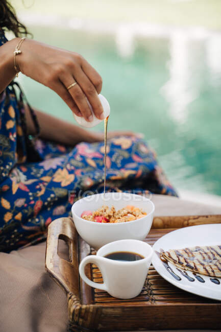 Crop unrecognizable female traveler pouring honey into power bowl with delicious breakfast on tray against coffee and crepe with chocolate sauce — Stock Photo
