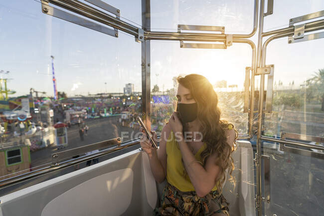 Женщина в защитной маске сидит в кабине колеса обозрения и просматривает мобильный телефон во время езды, развлекаясь на ярмарке во время коронавируса — стоковое фото