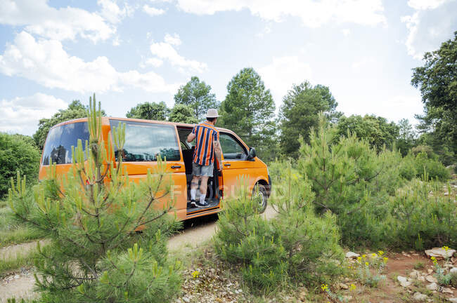 Vista posteriore di anonimo viaggiatore maschio in piedi con braccio alzato in furgone e godendo di viaggio su strada in estate nei boschi — Foto stock