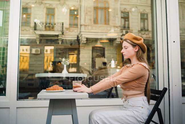 Trendy francese femmina in berretto seduta a tavola con gustoso caffè e dolce croissant durante la colazione in street cafè — Foto stock
