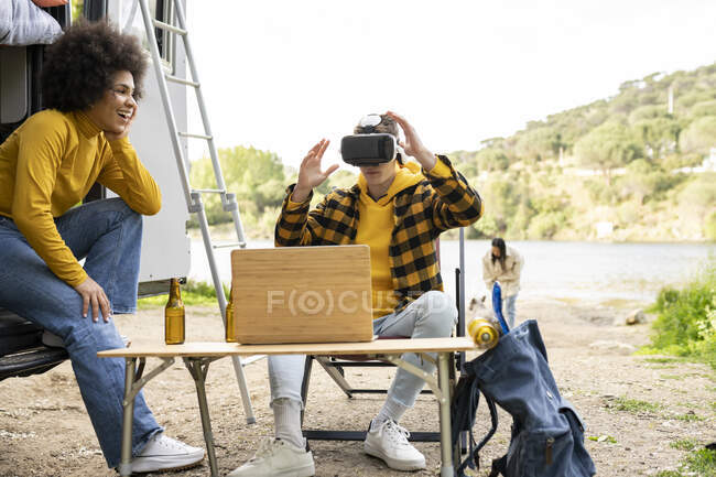 Fröhlicher Mann mit VR-Brille erkundet virtuelle Realität in der Nähe seiner schwarzen Freundin, während er gemeinsam in der Natur ruht — Stockfoto