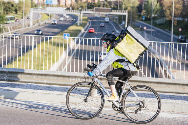 Vista laterale del corriere donna con borsa termica in sella alla bici sul ponte mentre consegna cibo in città — Foto stock
