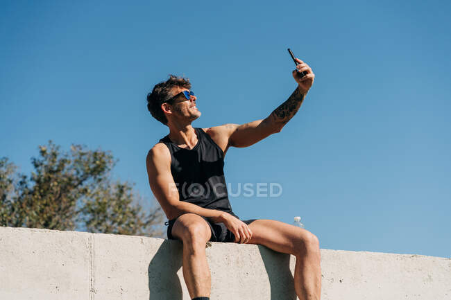 Ajuste atleta masculino em roupas esportivas tomando selfie com celular enquanto descansa na cerca após o treino — Fotografia de Stock