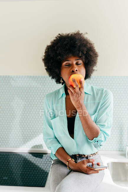 Moderna bella donna afroamericana con smartphone in mano seduta sul bancone della cucina con gli occhi chiusi a casa e mangiare mela — Foto stock