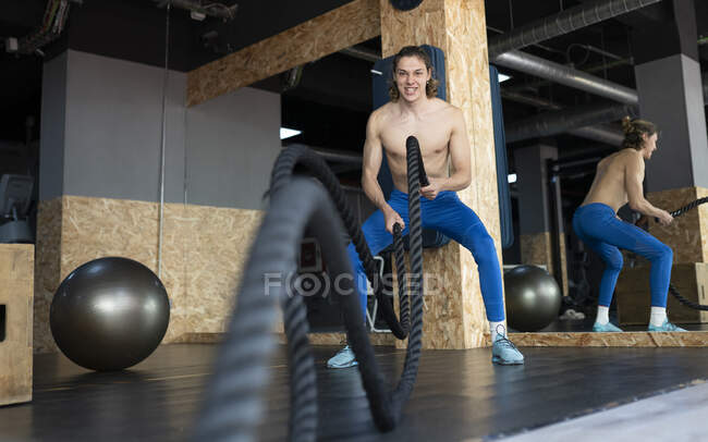 Мускулистый спортсмен с обнаженным туловищем, тренирующийся с боевой веревкой, глядя в камеру во время функциональной тренировки в гимназии — стоковое фото