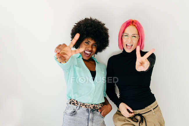 Modernos amigos femeninos multirraciales de moda en traje elegante gritando mientras mira a la cámara y mostrando v signo sobre fondo blanco - foto de stock