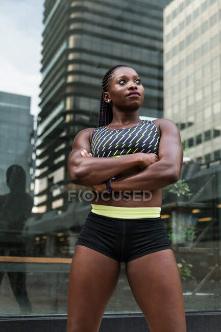 Femme noire positive en vêtements de sport gardant les bras croisés et regardant la caméra tout en se tenant debout sur un fond flou de la rue de la ville — Photo de stock