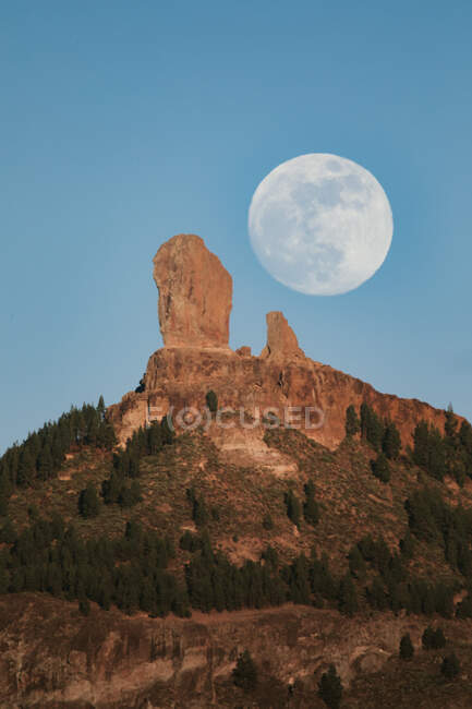 Spectacular landscape with large full moon in blue sky over rocky mountain peak with green forest in summer evening — Stock Photo