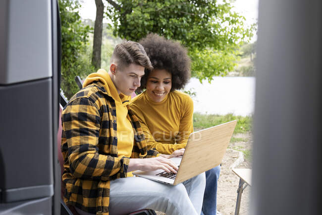 Jovem homem e mulher diverso em roupas casuais sorrindo e navegando netbook enquanto sentado perto de van durante a viagem na natureza — Fotografia de Stock