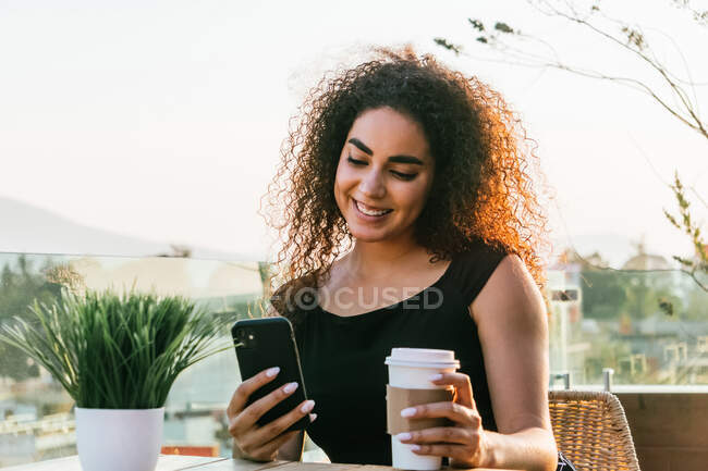 Fröhliche junge lockige hispanische Frau plaudert auf dem Handy, während sie Heißgetränk auf dem Imbissbecher trinkt und sich an sonnigen Sommerabenden auf der Caféterrasse ausruht — Stockfoto
